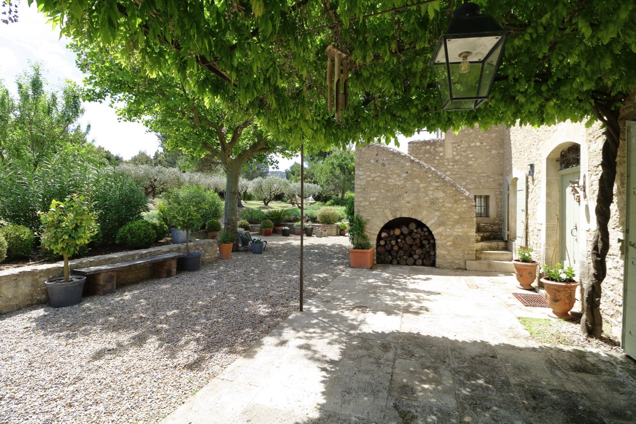 A tranquil courtyard with gravel floor, surrounded by lush green trees, featuring a traditional stone house with a wood log storage and potted plants. a lamppost stands in the center under the shade.